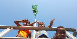 Looking up at 3 boys on EduMais's After-School Program at Leme fort who points towards the camera with Brazil flag and blue sky