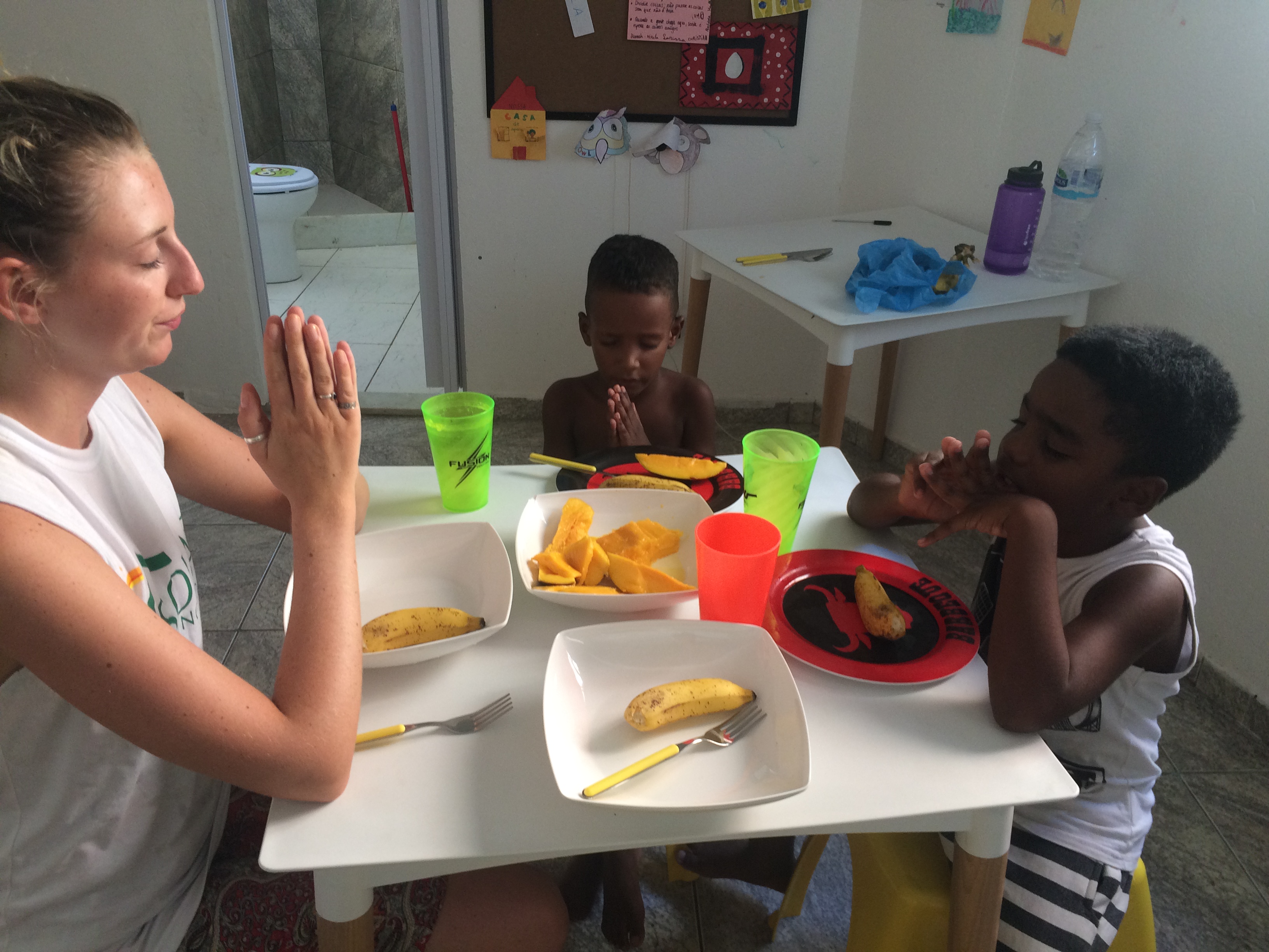 EduMais volunteer Eva and two boys on our after-school program give thanks before a snack of mango and bananas