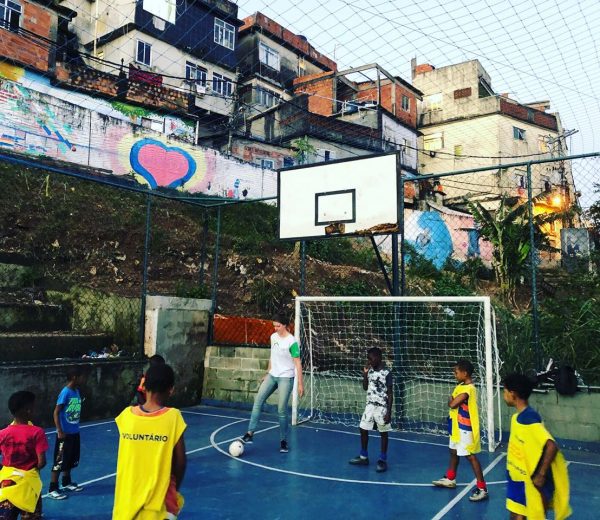 EduMais volunteer Iris plays with football with the kids on the pitch with houses of the Cantagalo favela visible in the background