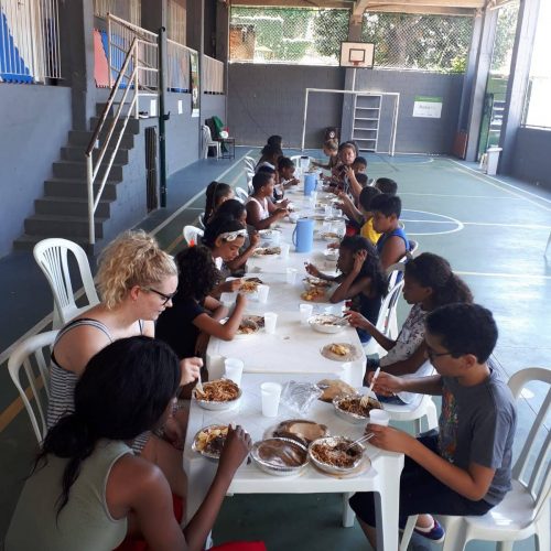 Students and volunteers at EduMais's English Summer Camp 2019 sit at long white tables having lunch