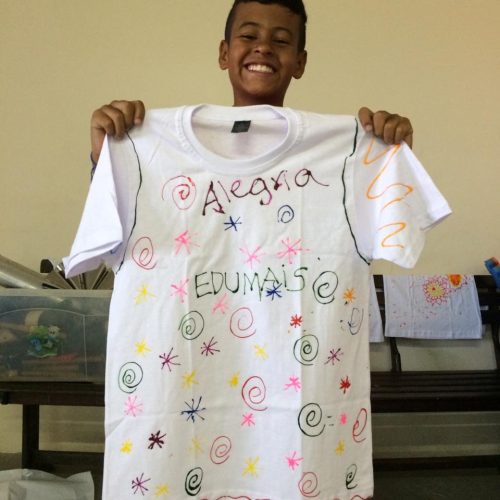 Student at EduMais's English Summer Camp holds up a t-shirt he designed with the word alegria (happiness)