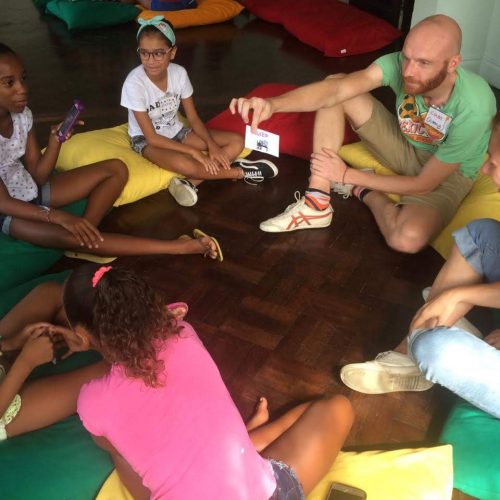 EduMais volunteer Coen holds up a flashcard for a group of students sat on colourful cushions at English Summer Camp 2019