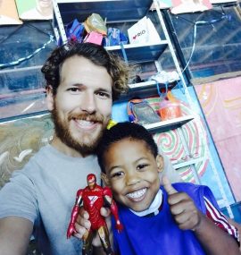 EduMais volunteer Tim with student making a thumbs up and holding an Iron Man toy